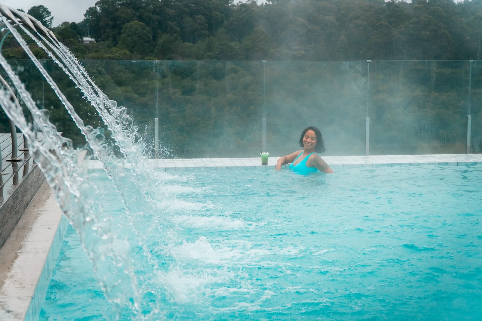 Piscina aquecida com jatos de água e vapor subindo, com a mata da Serra Gaúcha ao fundo em dia nublado