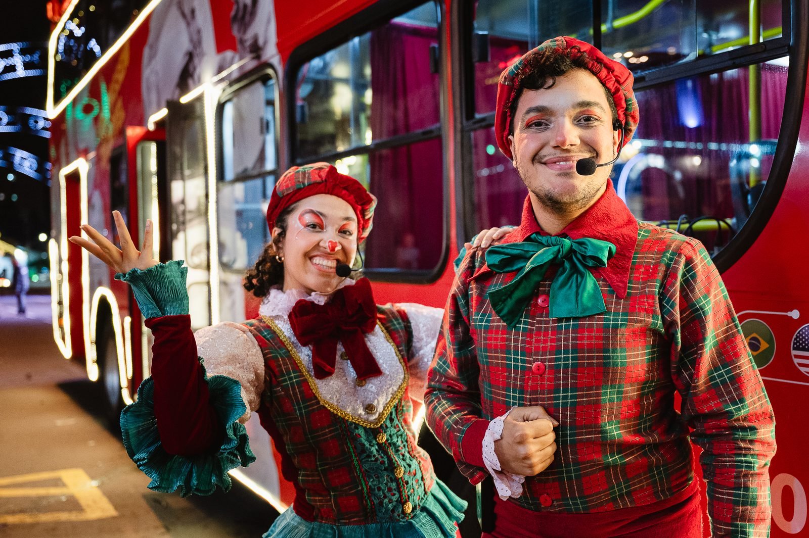 Casal de artistas em trajes xadrez de Natal ao lado do ônibus iluminado