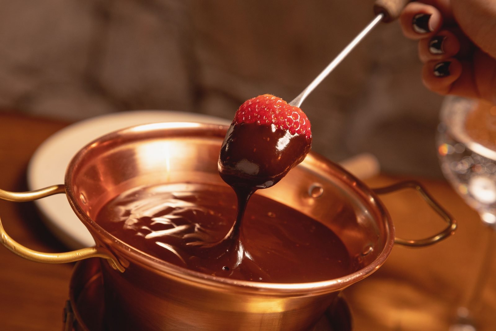 Panela de fondue sobre rechaud em mesa de madeira, com taça de vinho e prato de frutas