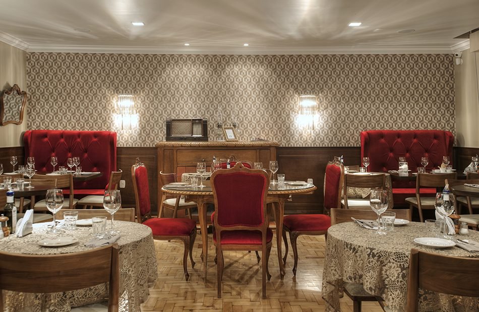 Salão do restaurante com poltronas vermelhas capitonê, mesas postas e papel de parede retrô