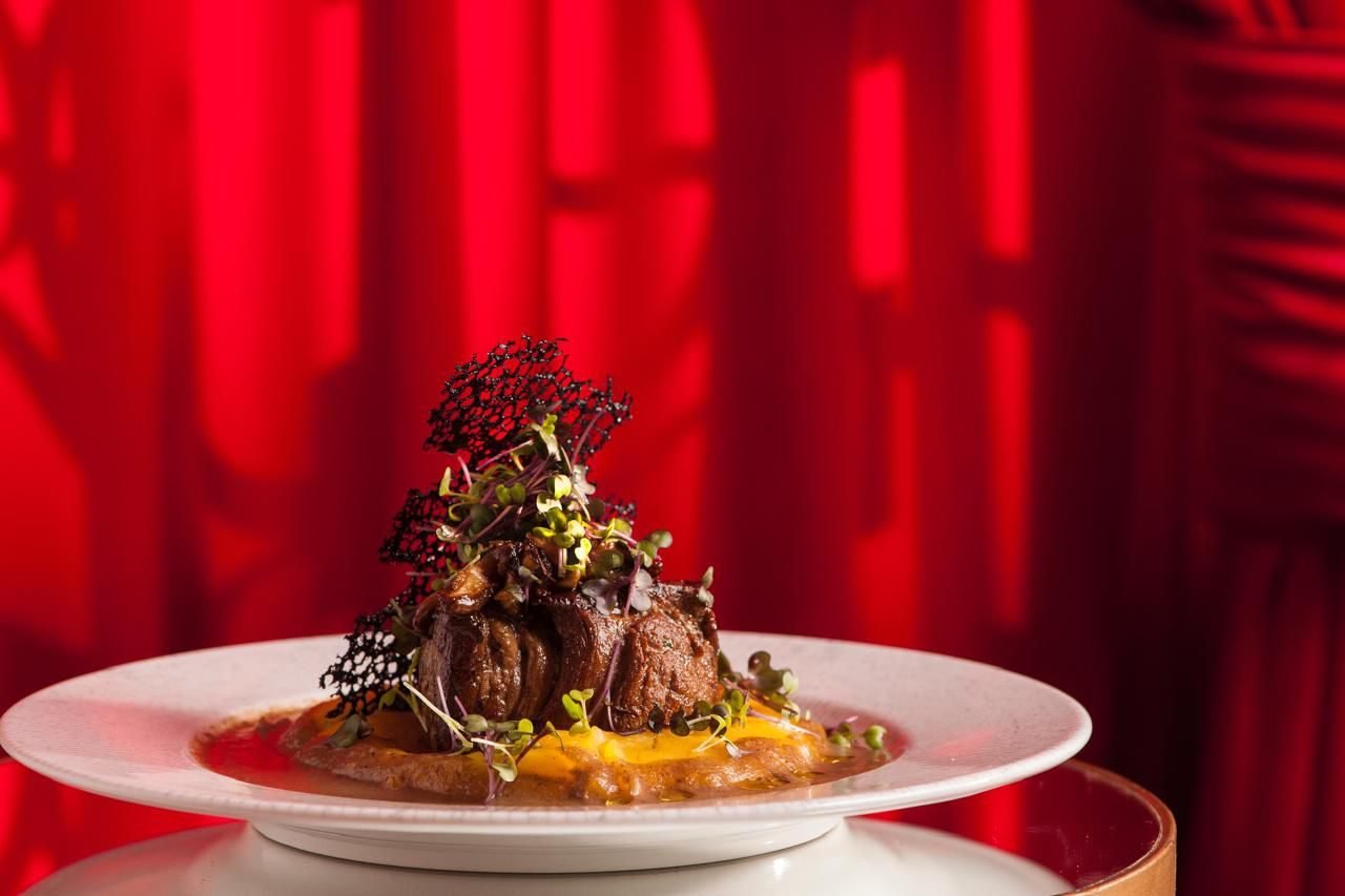 Medalhão de carne com molho e brotos servido em prato branco, com fundo vermelho