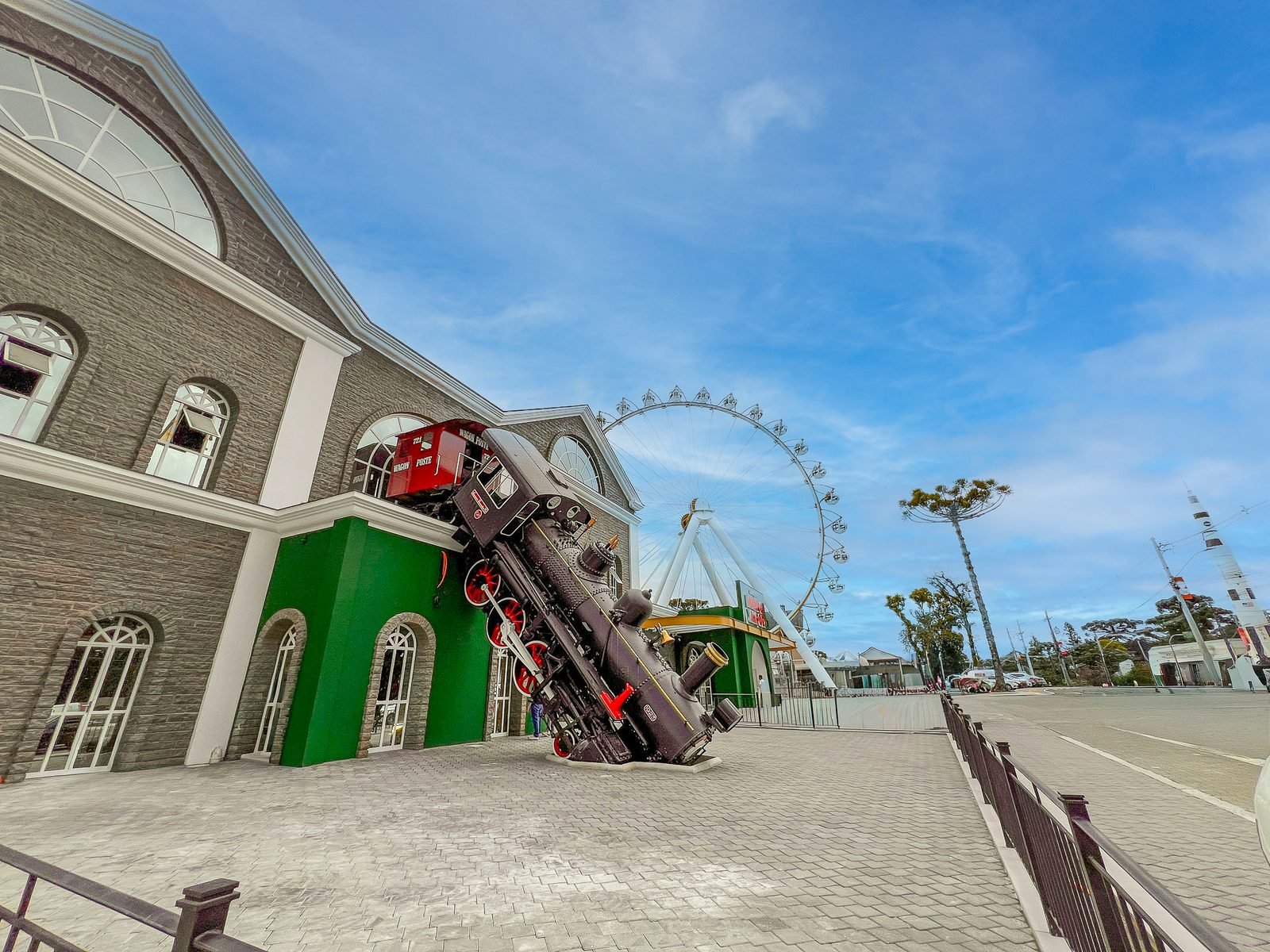 Locomotiva a vapor inclinada sobre a fachada do prédio do parque, com a roda gigante ao fundo