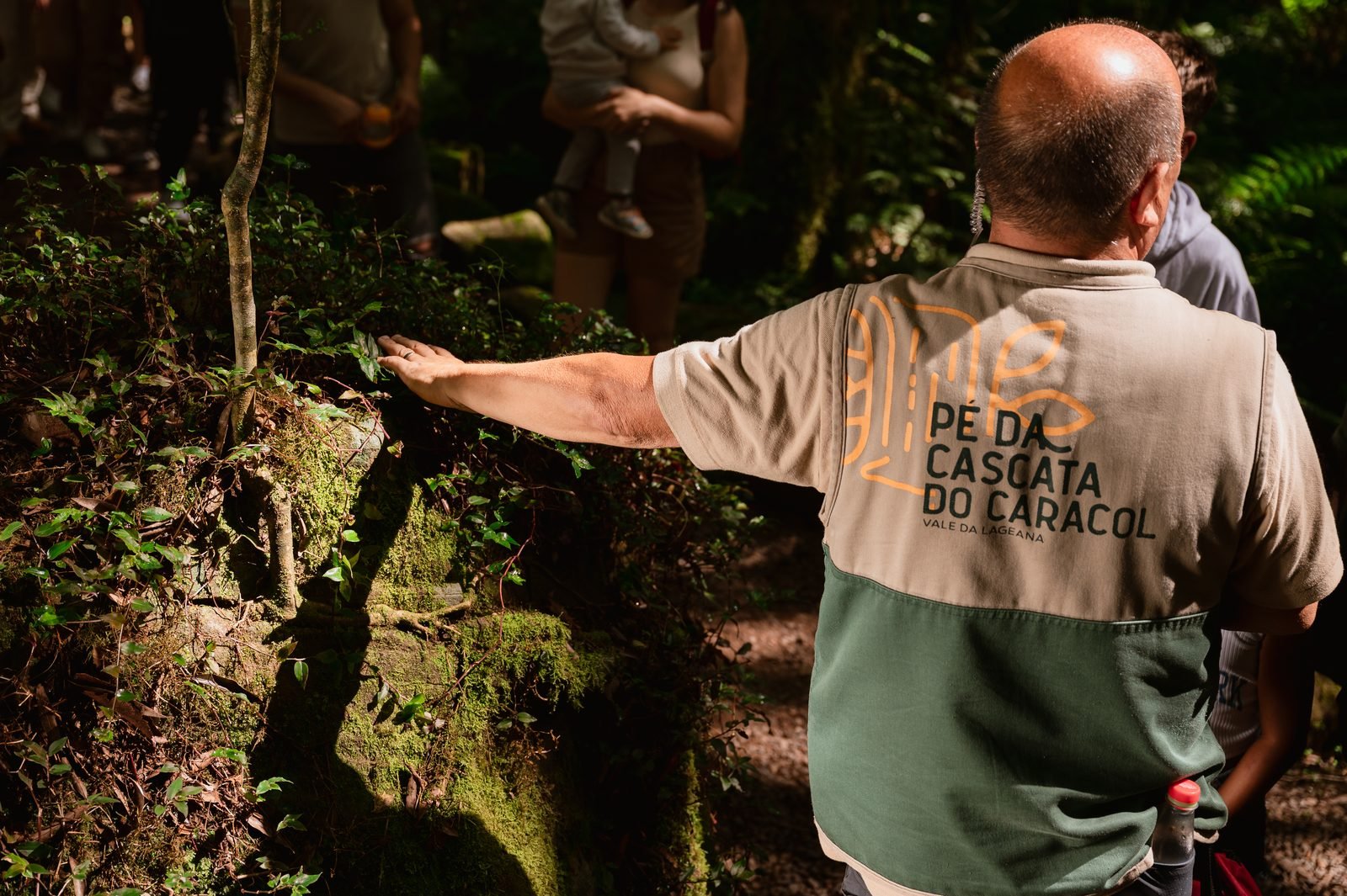 Condutor com colete do Pé da Cascata do Caracol apontando para a vegetação sobre uma rocha coberta de musgo