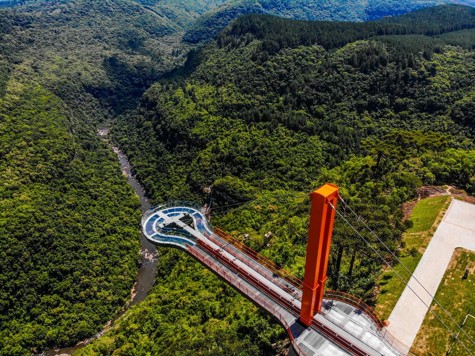 Vista aérea da plataforma circular de vidro do Skyglass avançando sobre o vale de mata, com o rio no fundo